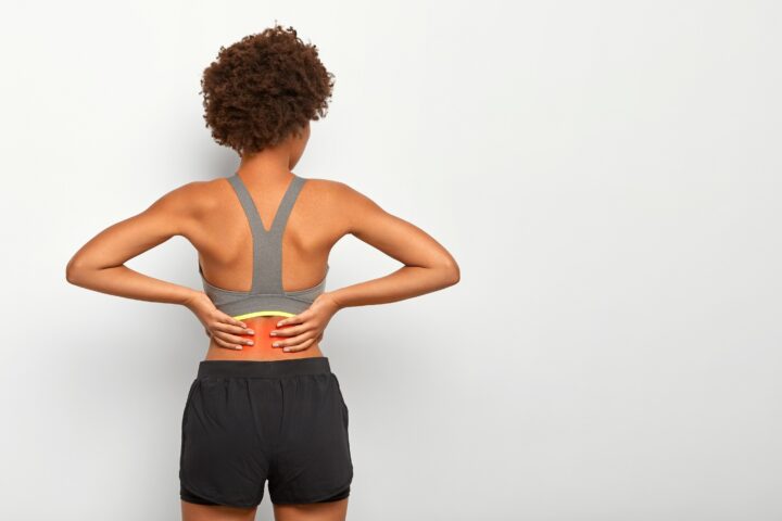 Photo of sporty woman with Afro haircut touches waist with both hands, feels pain in spine, shows location of inflammation, wears grey top, shorts, isolated over white wall. Lower back painful feeling